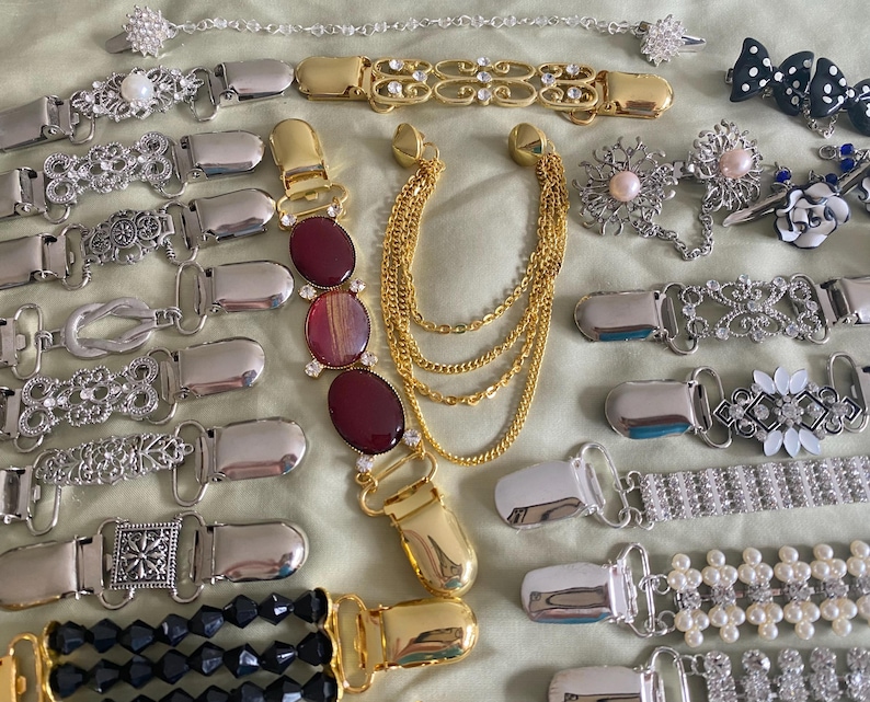May include: A collection of vintage belt clips and accessories in various designs. The clips feature silver and gold tones, with embellishments like rhinestones, pearls, and decorative patterns. Some clips have chain accents and oval-shaped red stones.