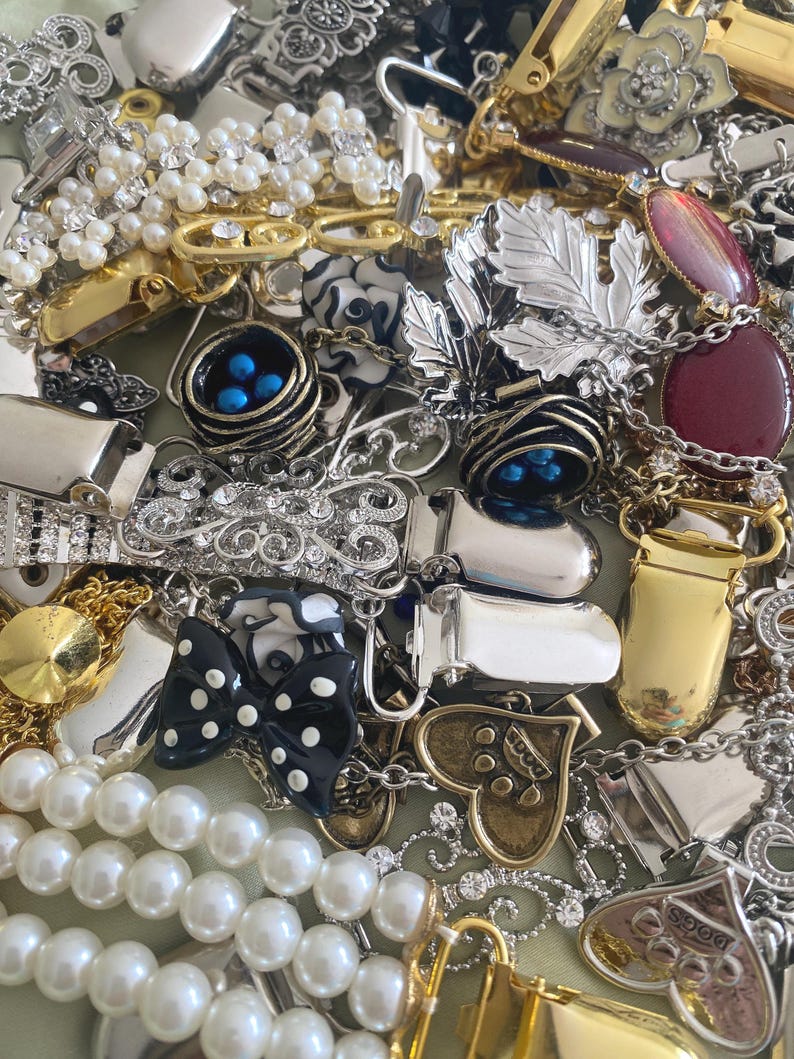May include: A collection of vintage jewelry pieces, including brooches, necklaces, and clips. The assortment features silver, gold, and pearl accents, with various designs such as leaves, bows, and hearts. Some pieces have embedded gemstones and enamel details.