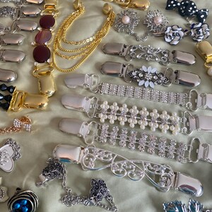 May include: A collection of various decorative sweater clips and brooches. The clips feature silver and gold metal finishes, adorned with rhinestones, pearls, and floral designs. Some clips have chain accents, and others feature gemstone embellishments. The brooches include pearl and floral designs.