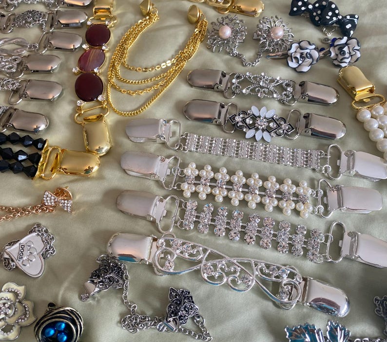 May include: A collection of various decorative sweater clips and brooches. The clips feature silver and gold metal finishes, adorned with rhinestones, pearls, and floral designs. Some clips have chain accents, and others feature gemstone embellishments. The brooches include pearl and floral designs.