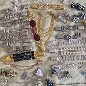 May include: A collection of various decorative suspender clips and chain accessories. The clips feature designs with rhinestones, pearls, and ornate metalwork in silver and gold tones. Some clips have chains with charms.