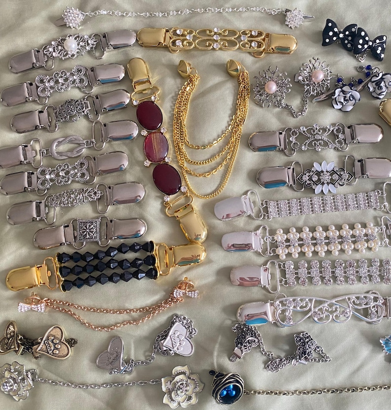 May include: A collection of various decorative suspender clips and chain accessories. The clips feature designs with rhinestones, pearls, and ornate metalwork in silver and gold tones. Some clips have chains with charms.