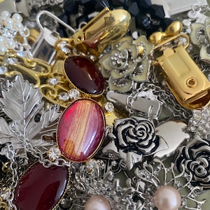 May include: A collection of vintage jewelry pieces, including brooches, earrings, and chains. The assortment features gold, silver, and black accents, with red and pink gemstones, pearls, and floral designs. Various textures and styles are visible.