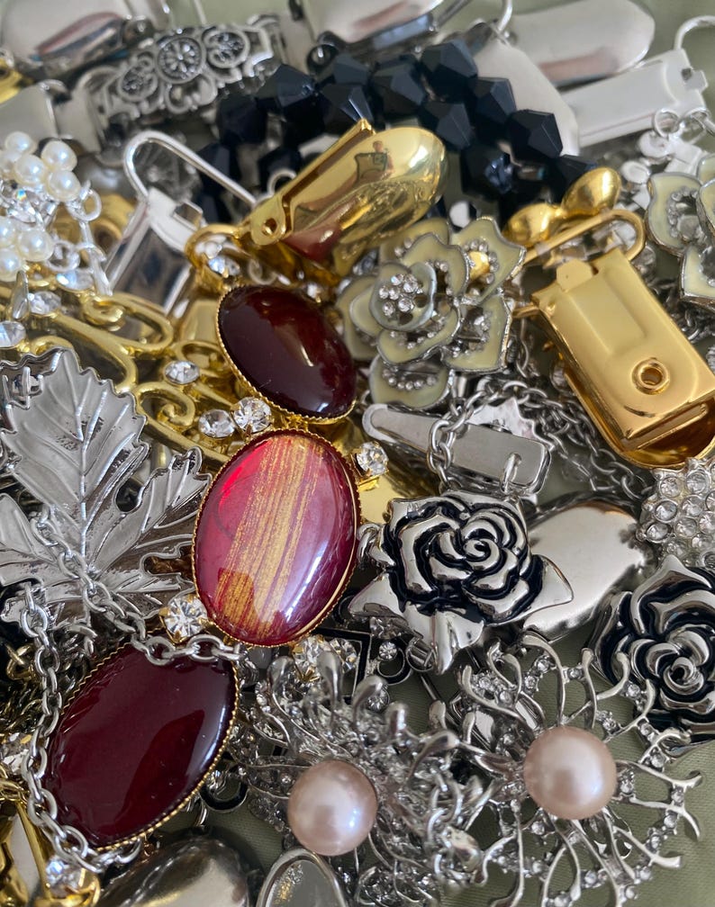 May include: A collection of vintage jewelry pieces, including brooches, earrings, and chains. The assortment features gold, silver, and black accents, with red and pink gemstones, pearls, and floral designs. Various textures and styles are visible.