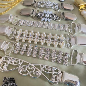 May include: An assortment of silver-toned sweater clips with various decorative elements. Designs include clear rhinestones, faux pearls, and floral patterns. The clips are displayed on a light green surface.