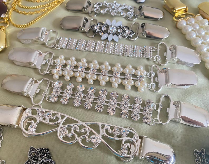 May include: An assortment of silver-toned sweater clips with various decorative elements. Designs include clear rhinestones, faux pearls, and floral patterns. The clips are displayed on a light green surface.