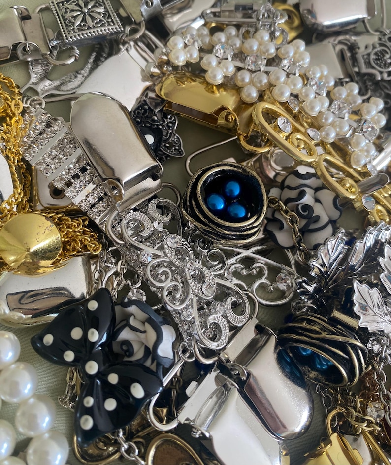 May include: A collection of vintage jewelry pieces, including brooches, necklaces, and earrings. The assortment features silver, gold, and pearl accents, with some pieces adorned with rhinestones and enamel details. A black and white bow-shaped brooch is also visible.