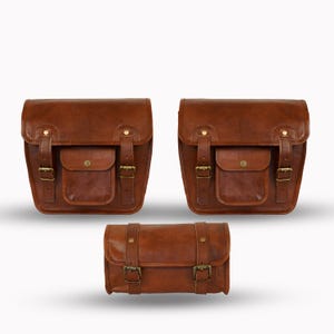 Pode incluir: Três alforjes de couro castanho com ferragens cor de latão. Duas bolsas maiores têm uma aba com fecho de fivela e um bolso frontal. A bolsa menor tem dois fechos de fivela e duas correias. Todas as bolsas são feitas de couro castanho.