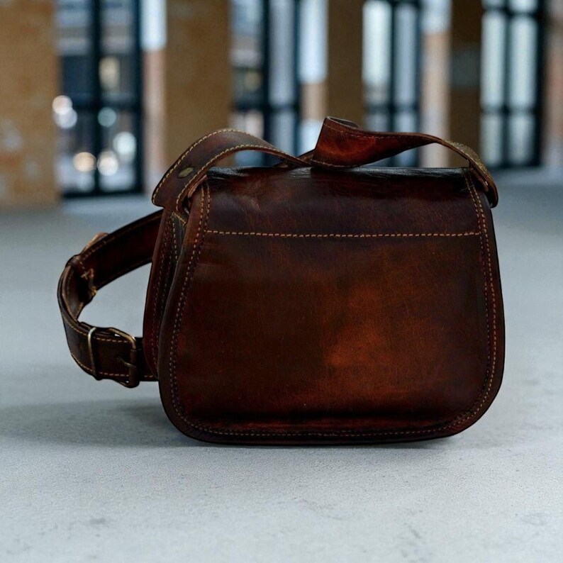 May include: A brown leather satchel bag with a shoulder strap and buckle fastening. The bag has a curved shape and visible stitching. The leather has a distressed appearance.