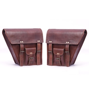 May include: Two brown leather motorcycle saddlebags. Each bag has a triangular shape, with a flap closure, two small pockets, and adjustable straps with buckles. The bags are made of a rich, dark brown leather.