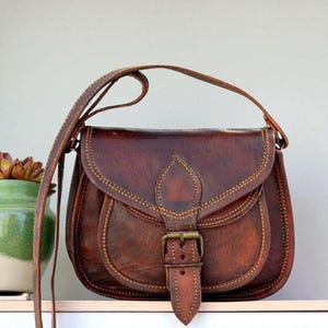 May include: A brown leather saddlebag with a long shoulder strap. The bag has a curved flap with a buckle closure and decorative stitching. The bag is on a white shelf. A green and white ceramic pot with a plant is to the left of the bag.
