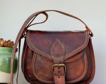 Könnte beinhalten: Eine braune Ledersatteltasche mit langem Schulterriemen. Die Tasche hat eine gebogene Klappe mit Schnallenverschluss und dekorativen Nähten. Die Tasche steht auf einem weißen Regal. Links von der Tasche befindet sich ein grün-weißer Keramiktopf mit einer Pflanze.