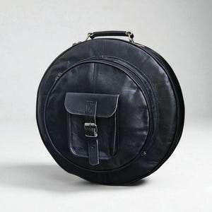 Op de afbeelding: Een zwarte leren instrumentenkoffer met een cirkelvormig ontwerp. Het heeft een handvat, een compartiment met ritssluiting en een voorvak met een gespsluiting. De koffer is ontworpen om muziekinstrumenten te beschermen en te vervoeren.