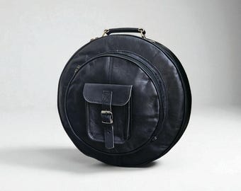 May include: A circular black leather instrument case. It has a handle, a zipped compartment, and a front pocket secured with a buckle. The case is designed for carrying and protecting musical instruments.