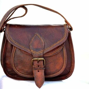 May include: A brown leather crossbody bag with a flap closure and adjustable strap. The bag features a buckle closure and decorative stitching. The bag is approximately 25 cm wide and 20 cm tall.