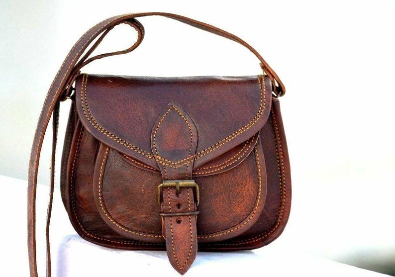 May include: A brown leather crossbody bag with a flap closure and adjustable strap. The bag features a buckle closure and decorative stitching. The bag is approximately 25 cm wide and 20 cm tall.