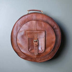 Puede incluir: Una bolsa redonda de cuero marrón con asa y un pequeño bolsillo delantero asegurado con una hebilla. La bolsa tiene un diseño circular y parece estar hecha de cuero de alta calidad. La bolsa está diseñada para transportar un instrumento musical.