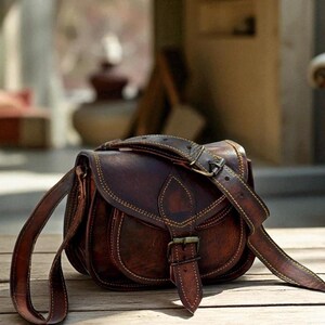May include: A brown leather satchel with a curved design and a long adjustable strap. The bag features a buckle closure and decorative stitching in a lighter brown colour. The bag is sitting on a wooden surface.