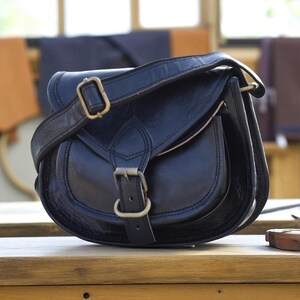 May include: A black leather saddle bag with a curved design. The bag has a flap closure with a buckle and an adjustable shoulder strap. The bag is on a wooden surface.