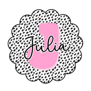 Puede incluir: Una letra "J" rosa con el nombre "Julia" en escritura cursiva, sobre un fondo blanco. La letra está dentro de un círculo con borde festoneado con un estampado dálmata en blanco y negro.