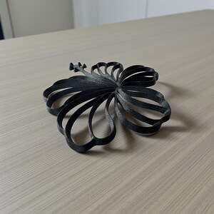 May include: A black, 3D-printed hibiscus flower, resting on a light wood-grain surface. The flower's petals are formed from curved, ribbon-like structures. The design is detailed, with a textured appearance, and the flower is approximately 6 inches in diameter.
