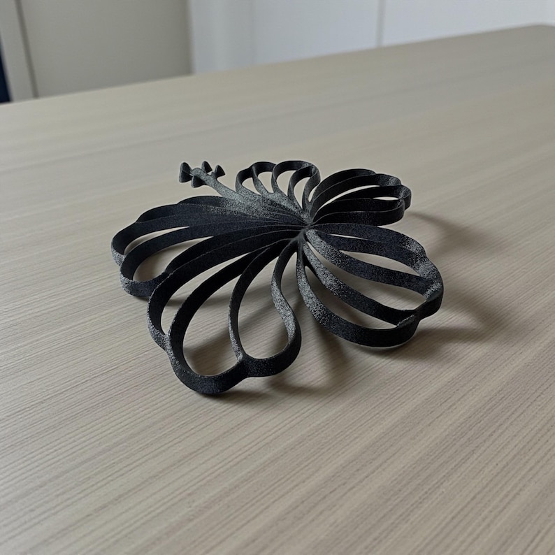May include: A black, 3D-printed hibiscus flower, resting on a light wood-grain surface. The flower's petals are formed from curved, ribbon-like structures. The design is detailed, with a textured appearance, and the flower is approximately 6 inches in diameter.