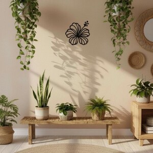 May include: A room with a neutral color palette, featuring various plants and decor. Two hanging plants in white pots flank a black hibiscus wall art. A wooden bench holds potted plants, and a round jute rug sits on the floor.