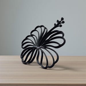 May include: A black, stylized hibiscus flower sculpture. The flower has multiple curved petals and a stem with small buds. The sculpture is placed on a light brown wooden surface against a gray background.