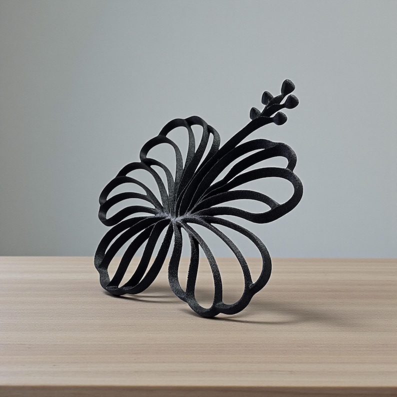 May include: A black, stylized hibiscus flower sculpture. The flower has multiple curved petals and a stem with small buds. The sculpture is placed on a light brown wooden surface against a gray background.