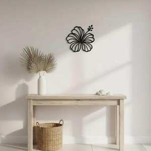 May include: A black hibiscus flower wall art decor. The flower is mounted on a white wall above a light wood console table. A white vase with dried palm leaves and a woven basket are also on the table.