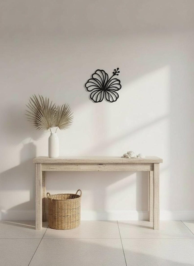 May include: A black hibiscus flower wall art decor. The flower is mounted on a white wall above a light wood console table. A white vase with dried palm leaves and a woven basket are also on the table.