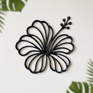 May include: Black hibiscus flower wall decor. The floral design is made of a solid black material. The flower has multiple petals and a stem with small buds. The background is white with green tropical leaves.