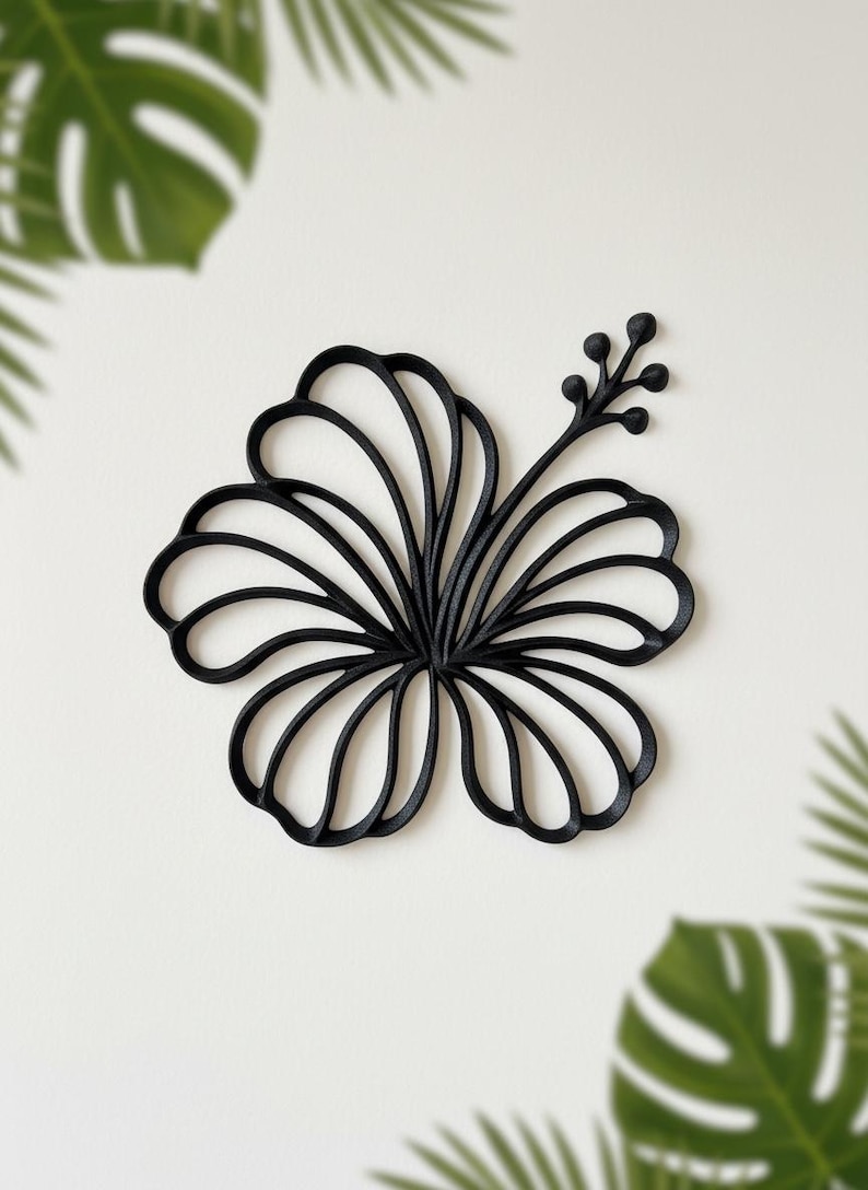 May include: Black hibiscus flower wall decor. The floral design is made of a solid black material. The flower has multiple petals and a stem with small buds. The background is white with green tropical leaves.