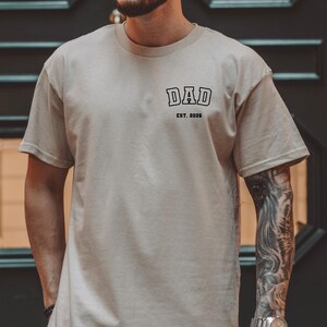 May include: A beige t-shirt with the word "DAD" printed in black, above "EST. 2026". The shirt has short sleeves and a crew neck. The person wearing the shirt has a tattoo on their arm and is wearing a watch.
