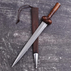 May include: A dagger with a polished steel blade and a dark wooden handle. The handle has a bulbous design. The dagger is sheathed in a brown leather scabbard with a silver tip and a leather cord.