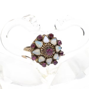 Puede incluir: Un anillo de oro con un grupo de ópalos blancos y piedras preciosas púrpuras engastadas en un diseño floral. El anillo se muestra en un cisne de cristal transparente.