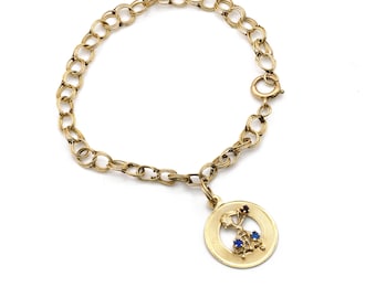 May include: Gold-toned charm bracelet with a round pendant. The pendant features a design with red and blue gemstones. The bracelet has a double-link chain and a clasp closure. A classic piece of jewelry.