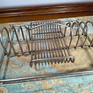 May include: A silver-colored metal toast rack with a rectangular base and multiple vertical dividers. The rack has a grid-like design for holding toast slices. The design includes rounded arches and decorative ball-shaped accents.
