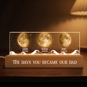 May include: A personalized night light featuring three illuminated moon phases with names and dates. The names David, Sophia, and Emily are displayed below each moon. The base of the light reads "THE DAYS YOU BECAME OUR DAD".