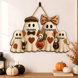 May include: A decorative wall hanging featuring four ghost figures labelled Emma, Grandpa, Grandma, and Noah. The ghosts have patches, a bow tie, a large floral bow, and carry a lantern and pumpkin buckets. It is suspended by a rope from a hook.