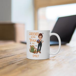 May include: White ceramic mug featuring a cartoon illustration of a gardener holding a basket of flowers. The mug has the text "Gardener of the Year" and the name "Emily" printed on it. The mug is on a wooden surface.