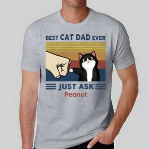 May include: Light gray t-shirt featuring a graphic with the text "BEST CAT DAD EVER" above an illustration of a black and white cat. The text "JUST ASK Peanut" is below the cat illustration.