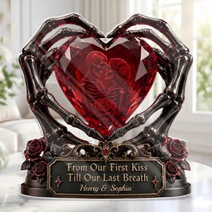 May include: A decorative sculpture featuring two dark skeletal hands forming a heart shape around a faceted red crystal heart. Inside the crystal is an etched image of two skeletons kissing with a rose. A base displays the text: From Our First Kiss Till Our Last Breath, Henry & Sophia, accented with sculpted red roses.