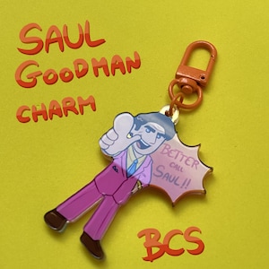 Pode incluir: Um chaveiro de acrílico com um personagem de desenho animado em um terno rosa apontando para frente, preso a um fecho lagosta laranja. O design inclui um balão de fala com o texto "BETTER CALL SAUL!!" e o personagem tem cabelo escuro.