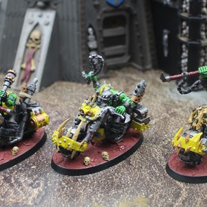 Puede incluir: Tres figuras en miniatura de orcos de piel verde montados en motocicletas amarillas y negras. Las motocicletas tienen extremos delanteros puntiagudos y están montadas sobre bases ovaladas. Los orcos sostienen armas y las bases están pintadas de rojo.