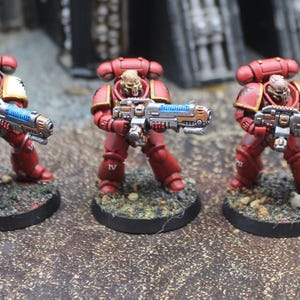 Puede incluir: Cinco figuras en miniatura con armadura roja, cada una sosteniendo un arma azul y plateada. Las figuras están sobre bases circulares negras. La armadura tiene detalles dorados. El fondo es borroso y de color neutro.