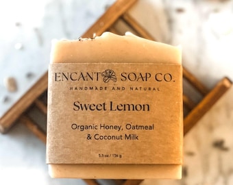 May include: A bar of handmade soap labeled "Sweet Lemon" by Encant Soap Co. The soap is light beige with a brown paper label. Ingredients include organic honey, oatmeal, and coconut milk. The bar weighs 5.5 oz / 156 g and rests on a wooden soap dish.
