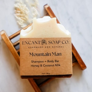 May include: A light-colored soap bar labeled "Mountain Man" sits on a wooden soap dish. The label reads "ENCANT SOAP CO." and lists ingredients: Honey & Coconut Milk. The bar is a shampoo and body bar, weighing 5 oz (142 g).