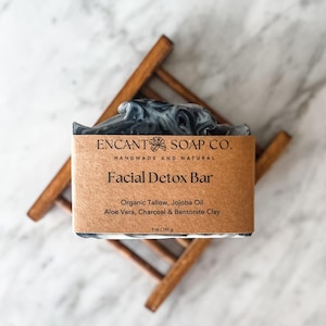 May include: A handmade facial detox bar with black and white marbling, resting on a small wooden rack. The bar is wrapped in a brown paper label that reads "ENCANT SOAP CO." and lists ingredients like organic tallow and jojoba oil. The bar weighs 5 oz / 142 g.