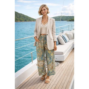 May include: A woman wearing a beige blazer, a white lace top, and patterned wide-leg pants. She is standing on a wooden deck with a blue ocean and green hills in the background. She is also wearing gold jewelry and sandals.